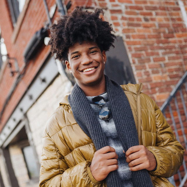 Young man wears mustard puffer jacket, gray scarf, and plaid shirt, standing outdoors near brick wall.