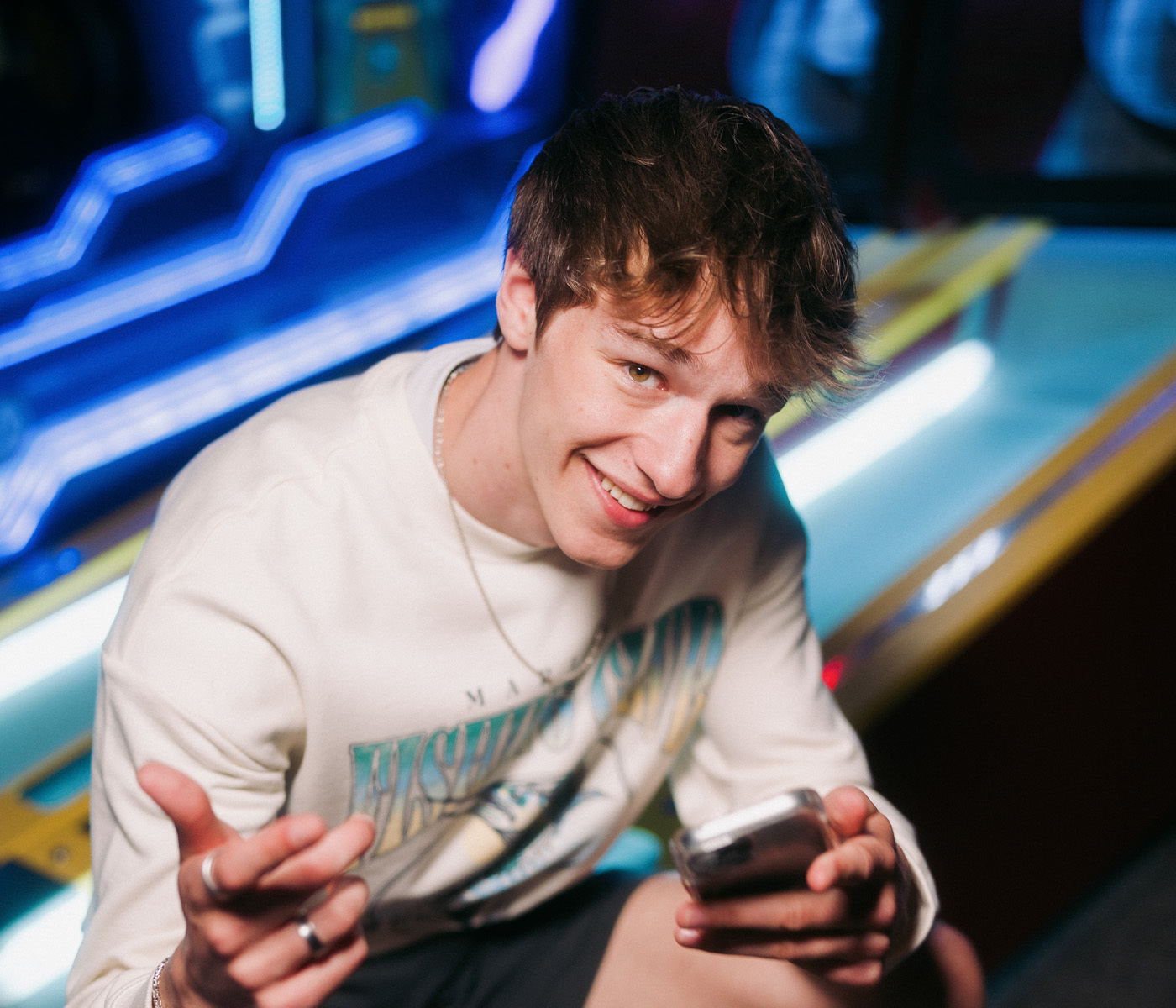 Person holding a smartphone while leaning toward the camera at an arcade with illuminated game lanes in the background.