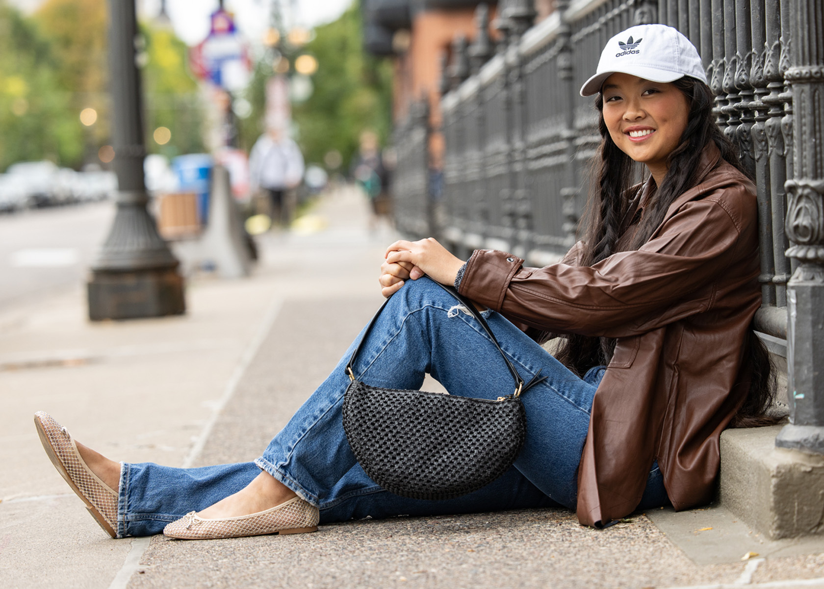 A young woman sitting on a sidewalk against a metal fence, wearing a brown leather jacket, blue jeans, beige flats, and a white cap, holding a black woven handbag.