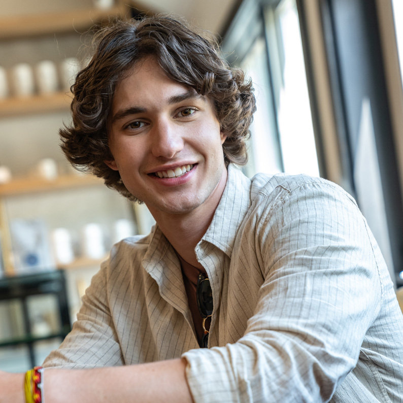 Person wearing a light beige striped button-up shirt with rolled sleeves, sitting indoors near a window with shelves in the background.