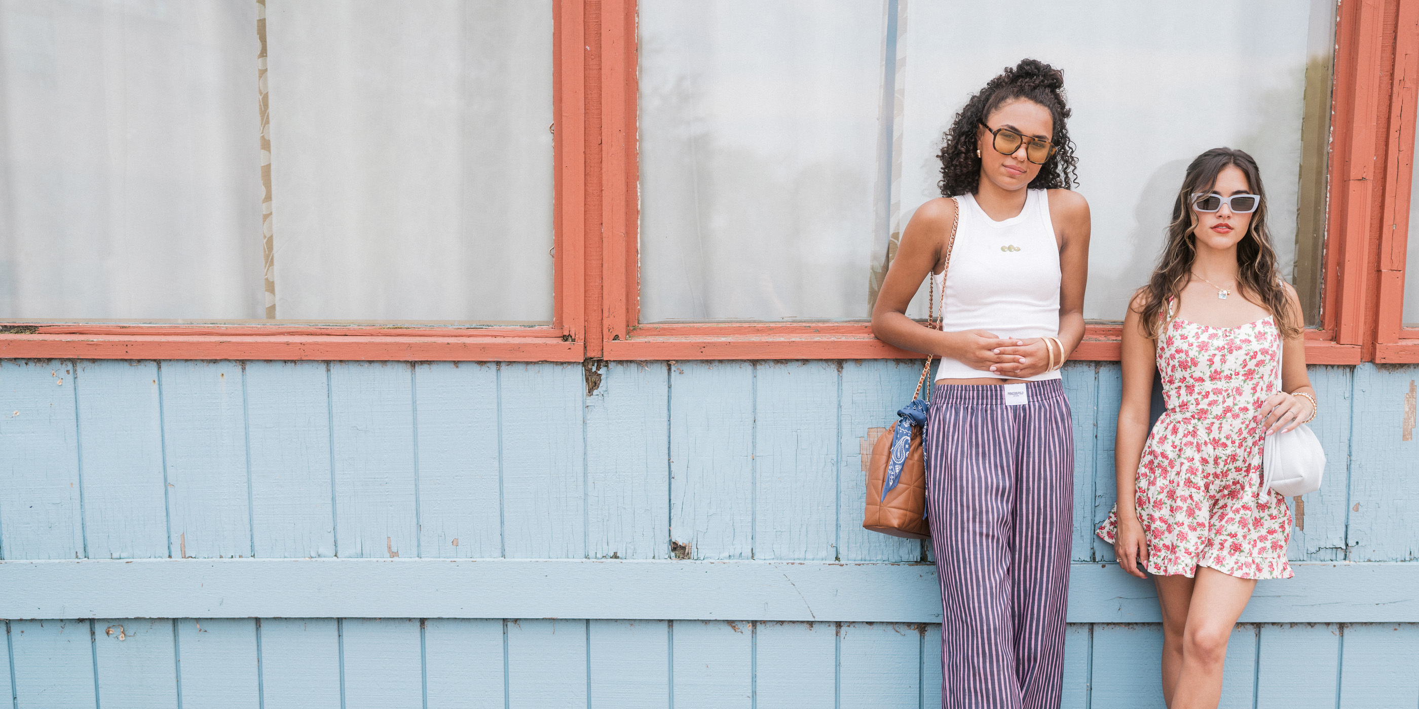 Two teens in fashionable clothing leaning against a wall with sunglasses on