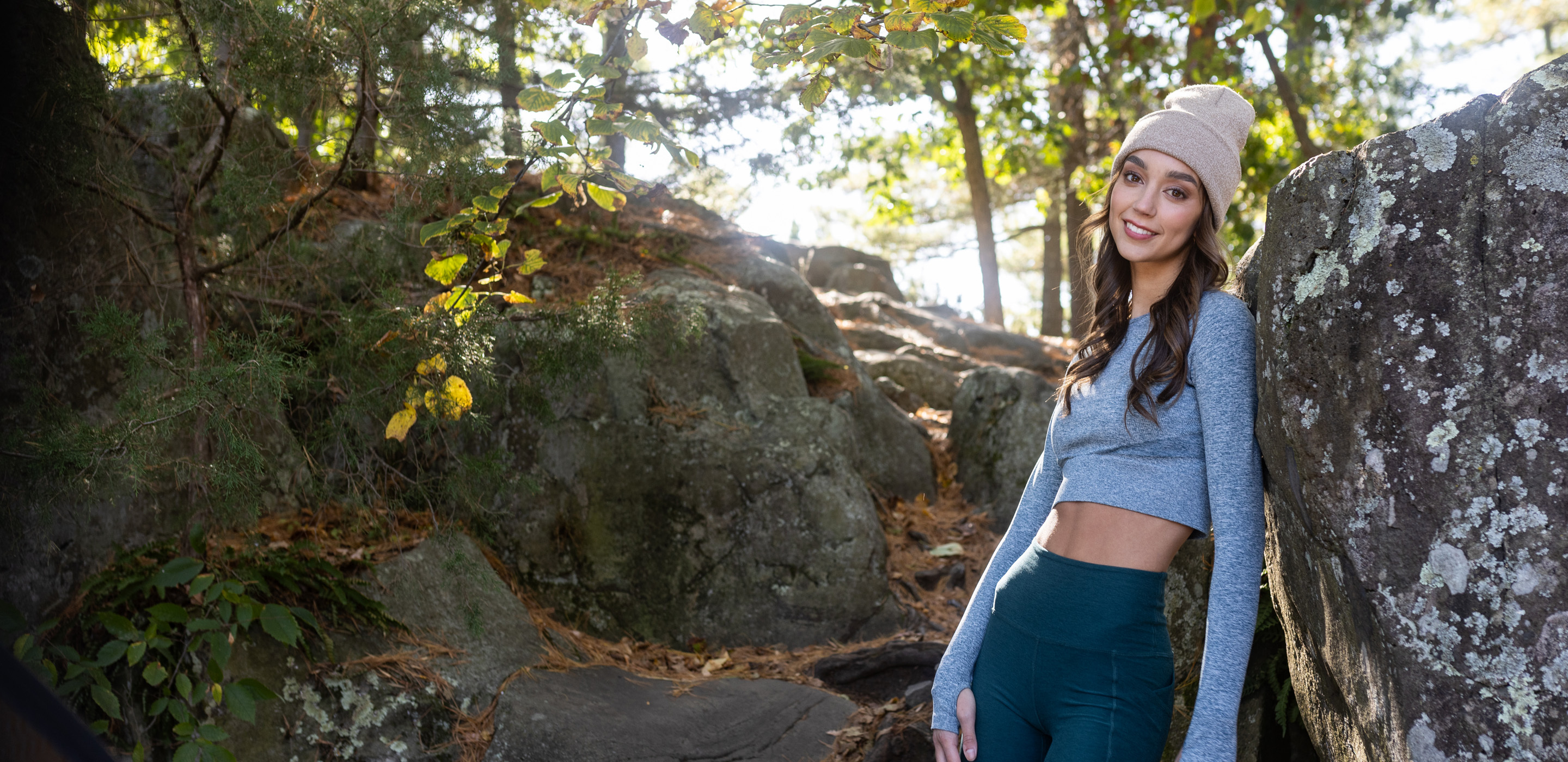A young woman wearing a gray long-sleeve crop top, teal leggings, and a beige beanie, standing among large rocks and trees outdoors.