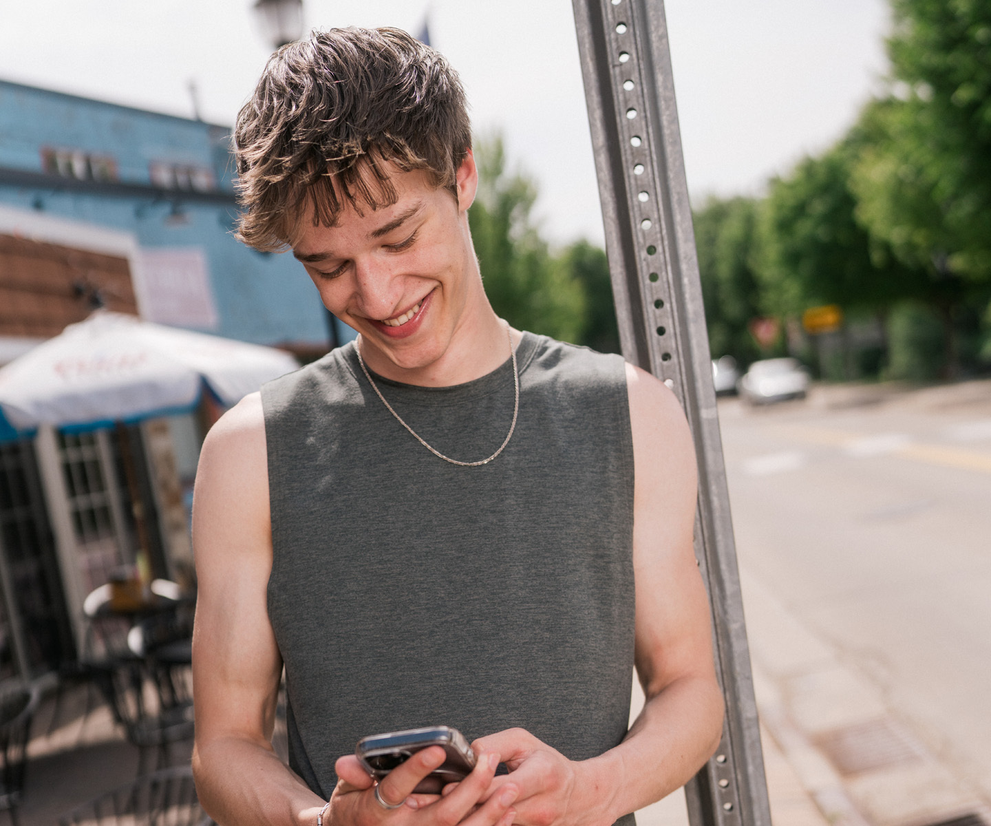 Person standing on a city sidewalk near a signpost, wearing a sleeveless gray top and holding a smartphone.