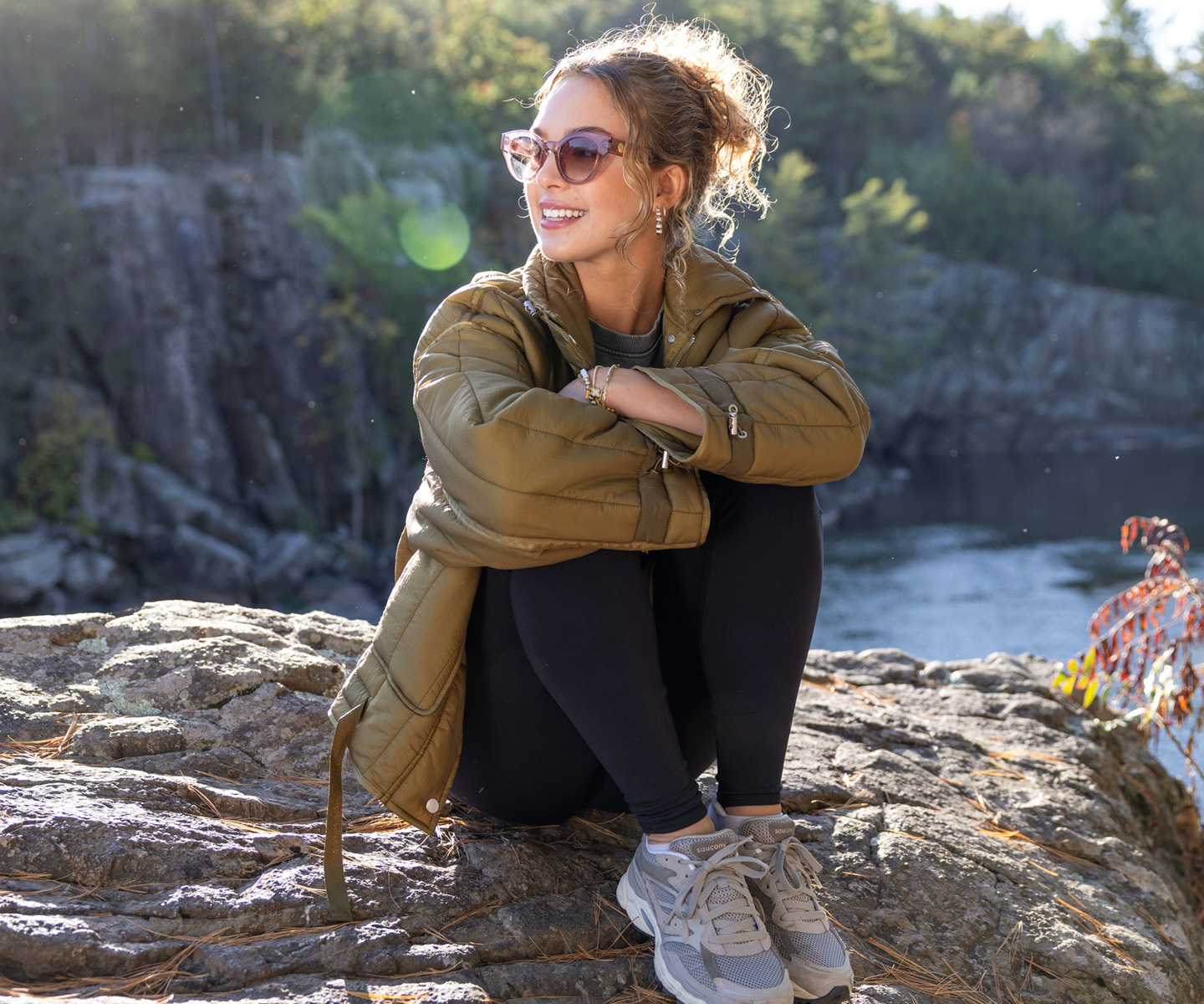 Woman sits on rocky riverbank, arms wrapped around knees, wearing olive quilted jacket, black leggings, and gray sneakers.