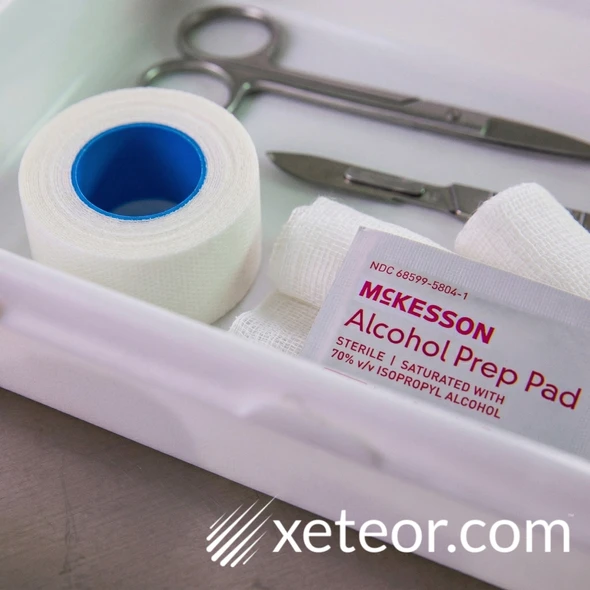 "A professional marketing photograph of a first-aid tray, showing a McKESSON Alcohol Prep Pad next to generic sterile gauze pads, a roll of medical tape with a plain blue core, medical scissors, and a scalpel, under a rose-pink banner that reads 'Professional first-aid supplies'