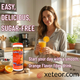 A marketing photograph for GoodSense Psyllium Fiber Supplement, showing a woman in her sun-drenched modern kitchen holding a glass of mixed orange fiber drink with an orange wedge on the rim. The bottle is completely isolated on the counter next to a bowl of yogurt and fruit and a measuring spoon with psyllium powder, all under warm morning sunlight.