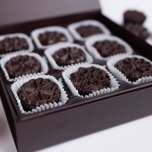 Close-up of a dark brown, rectangular gift box filled with small, individual chocolate treats. 

The chocolates are dark, rich brown, and each one is nestled in a small, white paper liner. The liners are slightly fluted/ridged, creating a subtle texture within the cavity.

The chocolates have a decorative pattern, a stylized snowflake-like shapes, pressed into the surface of the chocolate. They are arranged neatly in a grid pattern within the box.

The box itself is a dark, almost black brown colour, and appears to be made of a sturdy cardboard or similar material. The box's edges and the lid are sharply defined, creating a defined frame for the chocolates.

 A small portion of a separate, smaller dark-brown chocolate piece is visible in the upper-right corner of the image, outside the box, suggesting other chocolates or treats might be packaged elsewhere.

The background is a plain, white surface, which highlights the chocolate box and its contents. The lighting is bright and seems to be even, avoiding obvious shadows that might obscure the details of the chocolates.