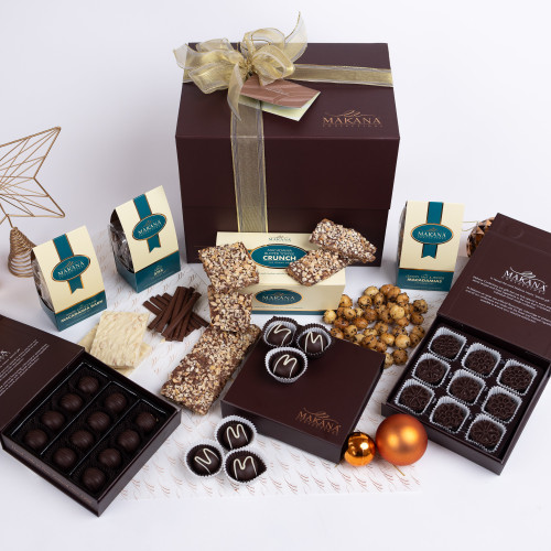 Full overhead shot of a lavish assortment of Makana chocolates and treats. 

A large, dark-brown gift box, tied with gold ribbon, forms the centrepiece.  Inside and around it are smaller boxes and containers containing various kinds of chocolates,  including truffles, bark, and other confections. 

Several small, rectangular boxes of Makana Crunch are visible, alongside a box of white chocolate pieces, and chocolate truffles in dark and milk chocolate in individual boxes. 

The chocolate treats are arranged in neat rows and clusters, with some individually wrapped in paper, and others in dark-brown cardboard boxes and tins. 

Scattered among the chocolates are a few pieces of Macadamia Butter Toffee Crunch. 

A gold-and-off-white patterned paper is beneath and amidst the various boxes and chocolates.

An orange-coloured Christmas ornament is placed on the paper next to the chocolates, and a gold star-shaped ornament is visible in the background to the left.

The lighting is bright and even, showcasing the details of the individual treats and the overall arrangement.
