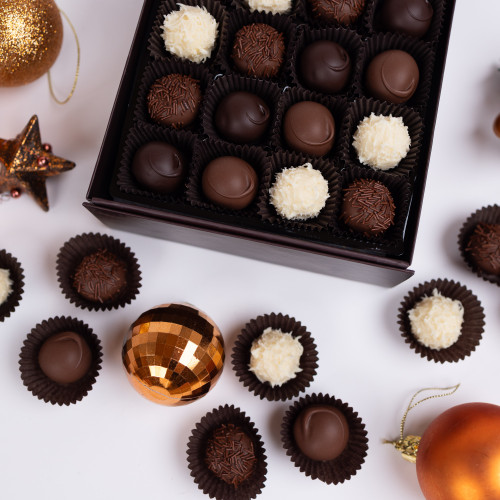 High-angle, full shot of a variety of chocolate truffles. 
A dark brown, rectangular gift box filled with individual chocolate truffles is centrally placed. The truffles are arranged in small, paper-lined compartments within the box, with a variety of dark chocolate, milk chocolate and white chocolate coated truffles.
Several individual, single-serving truffles in small, round, dark brown paper liners are scattered around the box. They create a visual array of the different truffle types and sizes around the box's edges. 
There are also two copper-coloured holiday ornaments, one resembling a disco ball, situated amongst the truffles on a white surface, adding a festive touch to the still life composition. A single, metallic orange-red holiday ornament hangs from a thin, gold string.
The background is plain white, which isolates the chocolates and ornaments for emphasis, drawing the viewer's eye to the assortment of treats. The lighting is even and bright, casting no significant shadows and allowing for detail visibility. High-angle, full shot of a variety of chocolate truffles. 
A dark brown, rectangular gift box filled with individual chocolate truffles is centrally placed. The truffles are arranged in small, paper-lined compartments within the box, with a variety of dark chocolate, milk chocolate and white chocolate coated truffles.
Several individual, single-serving truffles in small, round, dark brown paper liners are scattered around the box. They create a visual array of the different truffle types and sizes around the box's edges. 
There are also two copper-coloured holiday ornaments, one resembling a disco ball, situated amongst the truffles on a white surface, adding a festive touch to the still life composition. A single, metallic orange-red holiday ornament hangs from a thin, gold string.
The background is plain white, which isolates the chocolates and ornaments for emphasis, drawing the viewer's eye to the assortment of treats. The lighting is even and bright, casting no significant shadows and allowing for detail visibility.