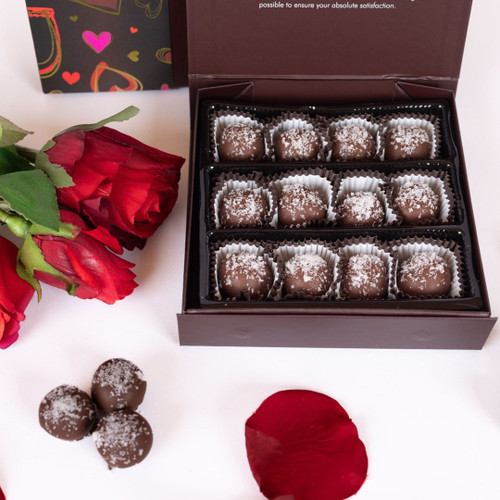 This visually appealing image captures an elegant arrangement ideal for romantic occasions. Central to the composition is a beautifully crafted box of chocolates, featuring two rows of individually wrapped truffles sprinkled with a light dusting of sea salt flakes, neatly positioned in paper liners. The rich, milk chocolate exterior glistens, inviting indulgence.

Surrounding the chocolate box are vibrant red roses, adding a touch of natural beauty and romance. The roses display lush petals and vivid green foliage, symbolizing love and affection. Scattered around the arrangement are delicate rose petals, creating a soft, romantic vibe. In the lower centre, a trio of chocolate truffles rests alongside additional rose petals, maintaining the colour theme.

The background is a crisp white, enhancing the richness of the chocolates and the bold red of the roses, making this image perfect for conveying love, celebration, or special occasions.