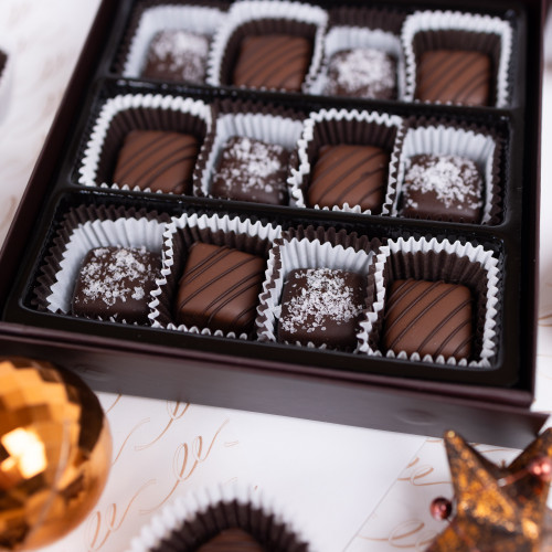 Close-up of a box of assorted chocolate truffles. 
The box is dark brown, rectangular, and has a slightly bevelled edge.  Individual chocolate dipped caramels are lined with white or brown, fluted paper liners. 
Within the compartments are various dark brown chocolate squares of differing designs.  The dark chocolate covered caramels have flecks of white sea salt on top, while the milk chocolate caramels have thin ribbons on dark chocolate decorating them.
The chocolates are arranged neatly in rows and columns within the box.
The box rests on a light-coloured surface, possibly paper or fabric with ornate script-like patterns in cream and/or beige.
Two copper-coloured Christmas ornaments are also visible in the foreground, partially visible and obscuring some of the script pattern. Also partially visible in the foreground is a portion of a bronze-coloured decorative item that resembles a crown or similar finial. Close-up of a box of assorted chocolate truffles. 
The box is dark brown, rectangular, and has a slightly bevelled edge.  Individual chocolate dipped caramels are lined with white or brown, fluted paper liners. 
Within the compartments are various dark brown chocolate squares of differing designs.  The dark chocolate covered caramels have flecks of white sea salt on top, while the milk chocolate caramels have thin ribbons on dark chocolate decorating them.
The chocolates are arranged neatly in rows and columns within the box.
The box rests on a light-coloured surface, possibly paper or fabric with ornate script-like patterns in cream and/or beige.
Two copper-coloured Christmas ornaments are also visible in the foreground, partially visible and obscuring some of the script pattern. Also partially visible in the foreground is a portion of a bronze-coloured decorative item that resembles a crown or similar finial.