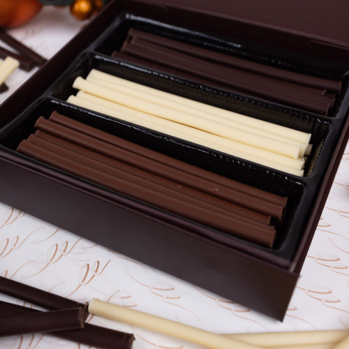 Close-up of a gift box containing chocolate sticks. 
The box is dark brown, leather-like, and has a slightly raised border around the edges. Inside, there are three compartments, each lined with black plastic. 
The compartments are filled with chocolate sticks, alternating colours of dark brown chocolate, light white and rich milk chocolate. The chocolate sticks are arranged neatly in each of the three sections.
 A white sheet of paper with a subtle, repeating pattern of elegant, stylized Makana swirl designs lies beneath and around the box. 
A few loose chocolate sticks are visible outside the box, on the white paper. Some scattered copper and brown-coloured Christmas decor items are visible in the background but blurred due to being out of focus. Close-up of a gift box containing chocolate sticks. 
The box is dark brown, leather-like, and has a slightly raised border around the edges. Inside, there are three compartments, each lined with black plastic. 
The compartments are filled with chocolate sticks, alternating colours of dark brown chocolate, light white and rich milk chocolate. The chocolate sticks are arranged neatly in each of the three sections.
 A white sheet of paper with a subtle, repeating pattern of elegant, stylized Makana swirl designs lies beneath and around the box. 
A few loose chocolate sticks are visible outside the box, on the white paper. Some scattered copper and brown-coloured Christmas decor items are visible in the background but blurred due to being out of focus.