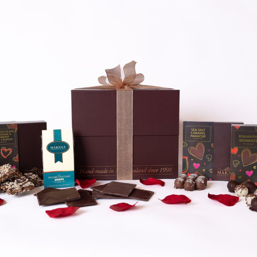 This image showcases a delightful assortment of luxurious chocolate treats, elegantly arranged for an appealing presentation. At the centre, a box labelled "Makana Confections" contains 72% dark chocolate snaps, highlighting its rich cocoa content and artisanal quality. Beside it, another package proudly displays “Dark Chocolate Macadamia & Cranberry Toffee Crunch,” capturing attention with its delightful flavour combination and inviting design.

Scattered around these treats are several pieces of the toffee crunch, showcasing a textured surface sprinkled with nuts and cranberries, and a selection of flat dark chocolate squares, suggesting indulgence in a variety of chocolate forms. Adding to the visual allure, vibrant red roses and scattered rose petals frame the arrangement, evoking a romantic and luxurious atmosphere, ideal for gifting or special occasions. The background features a chocolate-coloured box, emphasizing the handmade nature of the confections, making it a perfect treat for chocolate lovers.