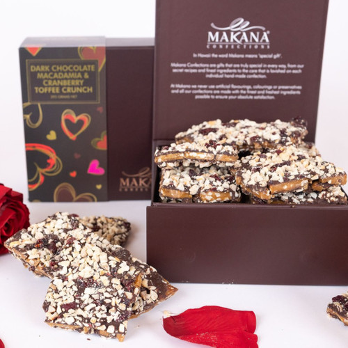 This image showcases an elegant arrangement of gourmet treats, featuring a box of "Dark Chocolate Macadamia & Cranberry Toffee Crunch" from Makana. The box, adorned with colourful heart motifs, hints at a special occasion or romantic gift. Inside, you can see the toffee crunch pieces, generously coated with dark chocolate and topped with crushed nuts and cranberries.

Surrounding the box are vibrant red roses, adding a touch of romance, while scattered rose petals enhance the overall romantic and indulgent atmosphere. The clean, white background makes the colours pop, drawing attention to both the luxurious chocolate treat and the beautiful flowers, creating an inviting presentation perfect for gifting or celebrating special moments.