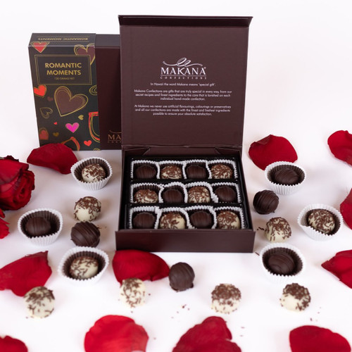 This image beautifully showcases a romantic gift arrangement, featuring an elegant chocolate box from Makana Confections, labeled "Romantic Moments." The box is open, revealing a delightful assortment of meticulously crafted chocolates, elegantly arranged within paper liners. Surrounding the box are scattered red rose petals and an arrangement of fresh red roses, enhancing the romantic ambiance.

In addition to the box of chocolates, individual chocolate truffles are artistically placed around, showcasing varieties that include dark and white chocolates, some decorated with chocolate shavings. The overall composition exudes a warm and inviting atmosphere, perfect for expressing love and affection. This image is ideal for use in promotional materials emphasizing romance, special occasions like anniversaries or Valentine's Day, or gourmet chocolate offerings.