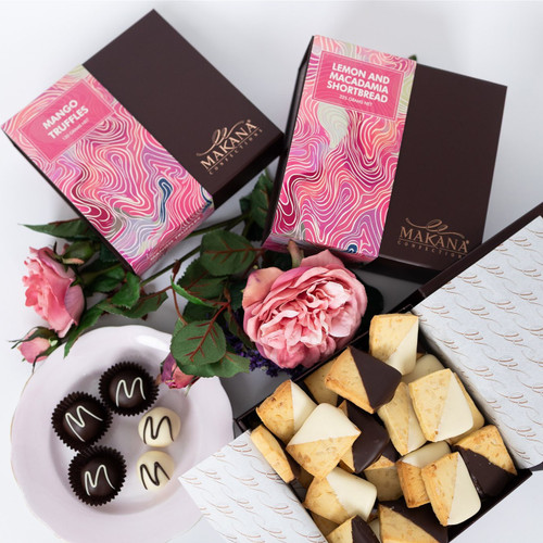 The picture showcases an elegant display of gourmet chocolates and cookies accompanied by fresh flowers, creating a sweet and inviting atmosphere.

In detail, the image features two beautifully designed boxes of confections from the brand "Makana." The box on the left is labelled "Mango Truffles," while the right box is for "Lemon and Macadamia Shortbread," both adorned with colourful, swirling patterns. Beneath the boxes lies a shallow pink plate holding four truffles: three dark chocolate ones with white swirls and one white chocolate truffle. 

Next to this, there's a larger open box filled with assorted shortbread cookies; some are dipped in dark and white chocolate, showcasing a variety of shapes and colors. Surrounding these treats are delicate pink roses and greenery, contributing a touch of elegance and a sense of warmth to the overall composition, evoking feelings of luxury and indulgence. The lighting is bright and soft, enhancing the appeal of the sweets and flowers.