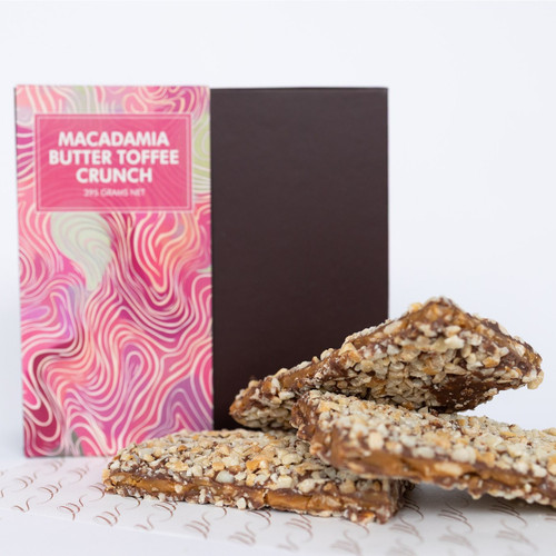 The picture features a box of macadamia butter toffee crunch candy alongside several pieces of the candy itself.

In detail, the image showcases a vibrant packaging design in shades of pink and green with swirling patterns, prominently featuring the text "MACADAMIA BUTTER TOFFEE CRUNCH" against a bold pink background. Behind the box is a dark brown panel, creating a nice contrast. In the foreground, three pieces of the toffee crunch are arranged, each topped with crushed macadamia nuts, giving them a textured appearance. The colors of the candies are a mix of golden brown and light beige, suggesting a rich, buttery flavor. The overall atmosphere of the picture is enticing and appetizing, with a clean, minimalist backdrop that emphasizes the sweetness and indulgence of the candy. The mood evokes a sense of delight and curiosity, inviting viewers to explore the treat further.