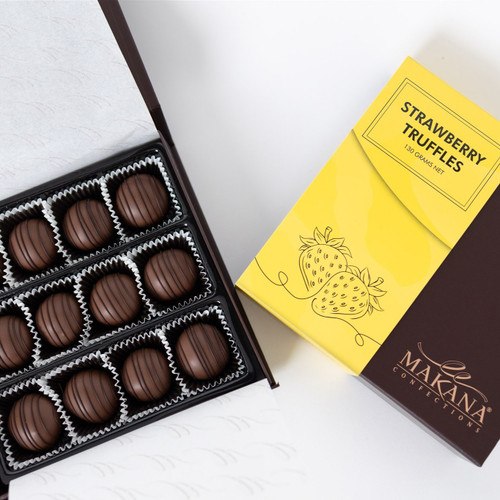High-angle, studio shot of a dark brown open box filled with nine chocolate truffles in white paper cups, viewed from top left. Next to it, is a yellow and brown box of strawberry truffles with strawberry graphic, and "Makana Confections" logo is printed in lower right. The backdrop is a clean white surface.