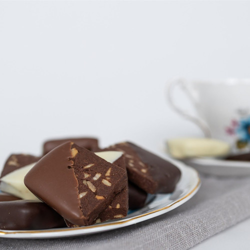 Close-up shot of chocolate dipped cocoa shortbreads sitting on a white decorative plate with a gold rim. The shortbreads are a chocolate colour dotted with nuts, with half of them dipped in dark chocolate and others dipped in white. A shallow depth of field places a teacup slightly out of focus in the background. A grey linen napkin rests underneath the plate, and the background is a clean white. The composition is well-lit, highlighting the shortbreads' textures.