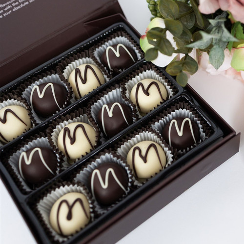 The picture features a beautifully arranged box of chocolates that exudes elegance and indulgence.

In detail, the image showcases a rectangular chocolate box with a dark brown exterior. Inside, there are two rows of twelve round chocolates, each placed in individual paper liners. The chocolates appear to be a mix of white and dark chocolate; some are coated in a rich dark chocolate while others are covered in creamy white chocolate, each adorned with a delicate swirl of contrasting chocolate on top. The background includes hints of soft greenery and pink flowers, suggesting a cheerful and inviting atmosphere. The overall mood of the picture is one of delight and luxury, making it feel celebratory and special, perfect for gifts or desserts.