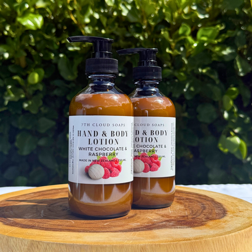 The image showcases two bottles of hand and body lotion, set against a backdrop of lush green foliage. The bottles are positioned on a circular wooden board, which is placed on a white surface, likely a table.
The bottles themselves are clear glass, revealing the lotion inside, which appears to be a warm, caramel-colored liquid. Each bottle is topped with a black pump dispenser. Attached to the front of each bottle is a white label, featuring the product's name and branding. The labels read "7TH CLOUD SOAPS HAND & BODY LOTION WHITE CHOCOLATE & RASPBERRY MADE IN NEW ZEALAND | 250 ML" and the other label reads "CLOUD SOAPS D & BODY OTION E CHOCOLATE & ASPBERRY NEW ZEALAND | 250 ML". A graphic of raspberries and a white chocolate truffle is displayed on the labels.
The wooden board has a natural, organic look with visible wood grain and a darker brown color. The background is a dense wall of green leaves, providing a natural and vibrant contrast to the bottles and the wooden board. The lighting appears to be natural, with soft shadows suggesting sunlight. The overall composition is well-balanced, with the bottles centered and the background providing a visual interest without distracting from the main subject. The image showcases two bottles of hand and body lotion, set against a backdrop of lush green foliage. The bottles are positioned on a circular wooden board, which is placed on a white surface, likely a table.
The bottles themselves are clear glass, revealing the lotion inside, which appears to be a warm, caramel-colored liquid. Each bottle is topped with a black pump dispenser. Attached to the front of each bottle is a white label, featuring the product's name and branding. The labels read "7TH CLOUD SOAPS HAND & BODY LOTION WHITE CHOCOLATE & RASPBERRY MADE IN NEW ZEALAND | 250 ML" and the other label reads "CLOUD SOAPS D & BODY OTION E CHOCOLATE & ASPBERRY NEW ZEALAND | 250 ML". A graphic of raspberries and a white chocolate truffle is displayed on the labels.
The wooden board has a natural, organic look with visible wood grain and a darker brown color. The background is a dense wall of green leaves, providing a natural and vibrant contrast to the bottles and the wooden board. The lighting appears to be natural, with soft shadows suggesting sunlight. The overall composition is well-balanced, with the bottles centered and the background providing a visual interest without distracting from the main subject.