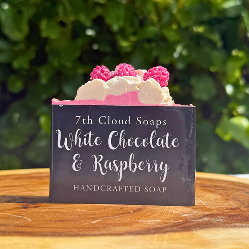 The image showcases a handcrafted soap bar presented on a wooden surface, likely a cutting board. The soap bar is rectangular and features several decorative elements. The top of the soap is adorned with what appear to be sculpted white and pink elements, resembling whipped cream and raspberries. Three raspberry-shaped decorations are prominently placed on top. The middle layer of the soap is a vibrant pink color, contrasting with the white and raspberry toppings. A black label is affixed to the front of the soap bar, displaying the words "7th Cloud Soaps," "White Chocolate & Raspberry," and "HANDCRAFTED SOAP" in a delicate, cursive font. The background of the image is a blurred green foliage, suggesting an outdoor setting. The lighting is soft and natural, enhancing the colors and textures of the soap. The wooden surface adds a rustic and natural feel to the composition. The overall tone of the image is inviting and aesthetically pleasing, emphasizing the handmade and gourmet nature of the soap. The image showcases a handcrafted soap bar presented on a wooden surface, likely a cutting board. The soap bar is rectangular and features several decorative elements. The top of the soap is adorned with what appear to be sculpted white and pink elements, resembling whipped cream and raspberries. Three raspberry-shaped decorations are prominently placed on top. The middle layer of the soap is a vibrant pink color, contrasting with the white and raspberry toppings. A black label is affixed to the front of the soap bar, displaying the words "7th Cloud Soaps," "White Chocolate & Raspberry," and "HANDCRAFTED SOAP" in a delicate, cursive font. The background of the image is a blurred green foliage, suggesting an outdoor setting. The lighting is soft and natural, enhancing the colors and textures of the soap. The wooden surface adds a rustic and natural feel to the composition. The overall tone of the image is inviting and aesthetically pleasing, emphasizing the handmade and gourmet nature of the soap.