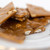 Close-up shot of maple-walnut brittle on a white plate. The lighting is soft, enhancing the caramel colour and nut textures. The brittle itself is arranged in irregular pieces, stacked upon each other, drawing the eye to the centre of the frame. The shallow depth of field puts emphasis on the brittle itself.