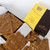 High angle shot of a partially opened box of Maple & Walnut Brittle with brittle pieces scattered around the box on a patterned white surface. The box is split diagonally between yellow (top left) with black text & a maple leaf/walnut illustration and dark brown (bottom right) with the Makana logo in silver. The brittle pieces are various shades of brown and contain visible walnuts. The background is a soft white. The composition focuses on the textures of the brittle and the contrast between the box colours.
