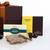 Studio shot of Makana confectionary products arranged on a white surface. A large, partially visible gift box fills the upper left, partly obscuring similarly coloured boxes behind the toffee and truffle boxes. Focus is on the Makana Macadamia Butter Toffee Crunch box in the centre. The toffee pieces create visual texture, drawing the eye. Lighting is soft and diffused, highlighting the colours and textures of the confectionery and packaging. The composition suggests a sense of luxury and indulgence.