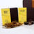 Studio shot displaying two Makana candy boxes: a yellow box with "MAPLE & WALNUT BRITTLE 395 GRAMS NET" in dark letters with Maple leaf and walnut images, and another yellow box with a black bunny image that reads "HOT CROSS BON BONS 130 GRAMS NET." The brittle candy pieces are piled on a plate, with Makana logo text appearing subtly along with "Happy Easter". The chocolate eggs and boxes are placed on a surface with a repeating pattern. The background extends with stacked boxes, lit in soft lighting.