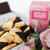 The picture showcases a tempting assortment of shortbread cookies alongside a colourful packaging box.

In detail, the image features an appealing arrangement of shortbread cookies that are rectangular in shape, some coated in dark chocolate and others coated in white chocolate. The shortbreads have a golden-brown hue with visible specks of macadamia nuts and lemon zest, suggesting a delightful flavour profile. Next to the cookies is a vibrantly designed box labelled "Lemon and Macadamia Shortbread," which presents a swirled pink and purple pattern that adds a playful element to the composition. Surrounding the cookies are soft, muted flowers and greenery, enhancing the overall aesthetic and creating a sweet, inviting atmosphere. The mood of the picture is warm and cheerful, evoking a sense of indulgence and enjoyment, perfect for gifting or special occasions.