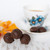 Close-up, medium shot of chocolate truffles in the foreground, arranged in a small pile on a white surface with light brown swirls. The background is softly blurred, featuring a teacup and saucer with blue floral designs and a pale beverage. The composition creates depth through selective focus, highlighting contrast between the richness of the chocolate and the delicacy of the tea ware. Bright white background dominating overall. A partial orange flower is seen in the left side of the picture, only slightly in focus.