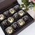The picture features a beautifully arranged box of chocolates that exudes elegance and indulgence.

In detail, the image showcases a rectangular chocolate box with a dark brown exterior. Inside, there are two rows of twelve round chocolates, each placed in individual paper liners. The chocolates appear to be a mix of white and dark chocolate; some are coated in a rich dark chocolate while others are covered in creamy white chocolate, each adorned with a delicate swirl of contrasting chocolate on top. The background includes hints of soft greenery and pink flowers, suggesting a cheerful and inviting atmosphere. The overall mood of the picture is one of delight and luxury, making it feel celebratory and special, perfect for gifts or desserts.