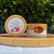 The image showcases two containers of hand and body butter displayed on a wooden serving board. The background is blurred, featuring lush green foliage, suggesting an outdoor setting. The wooden board, with its natural grain and warm tones, provides a rustic and organic feel to the composition.
The first container, a large, circular jar, is positioned on the left. Its wooden lid is prominently visible, with a label affixed on top. The label reads "7TH CLOUD SOAPS" at the top, followed by "HAND & BODY BUTTER," "WHITE CHOCOLATE & RASPBERRY" and "FOR SENSITIVE SKIN | MADE IN NZ | 100ML." A graphic of raspberries and a white chocolate ball is also included on the label, illustrating the product's flavor profile.
The second container, located to the right, is a smaller, frosted glass jar. It also has a wooden lid, with an oval-shaped label that reads "le MAKANA" in an elegant font. The jar contains a white, creamy substance, presumably the hand and body butter itself.
The products are arranged on a flat surface, likely a white tablecloth, which contrasts with the wood and the greenery in the background. The lighting appears soft and natural, enhancing the colors of the products and the wooden elements. The composition is well-balanced, with the containers placed at different angles, creating visual interest. The overall impression is one of natural beauty, quality ingredients, and a focus on skincare. The image showcases two containers of hand and body butter displayed on a wooden serving board. The background is blurred, featuring lush green foliage, suggesting an outdoor setting. The wooden board, with its natural grain and warm tones, provides a rustic and organic feel to the composition.
The first container, a large, circular jar, is positioned on the left. Its wooden lid is prominently visible, with a label affixed on top. The label reads "7TH CLOUD SOAPS" at the top, followed by "HAND & BODY BUTTER," "WHITE CHOCOLATE & RASPBERRY" and "FOR SENSITIVE SKIN | MADE IN NZ | 100ML." A graphic of raspberries and a white chocolate ball is also included on the label, illustrating the product's flavor profile.
The second container, located to the right, is a smaller, frosted glass jar. It also has a wooden lid, with an oval-shaped label that reads "le MAKANA" in an elegant font. The jar contains a white, creamy substance, presumably the hand and body butter itself.
The products are arranged on a flat surface, likely a white tablecloth, which contrasts with the wood and the greenery in the background. The lighting appears soft and natural, enhancing the colors of the products and the wooden elements. The composition is well-balanced, with the containers placed at different angles, creating visual interest. The overall impression is one of natural beauty, quality ingredients, and a focus on skincare.