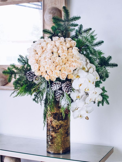 This absolutely majestic floral arrangement is sure to capture your attention with its grandiose design and crisp winter whites. Seventy-five ivory roses draw the eye next to a waterfall of cascading orchids. Snow flocked Christmas greens and pinecones provide an element of the wild. This breathtaking design rests atop a 22" by 8" birchwood lined glass vase and stands at an impressive 45" tall overall.