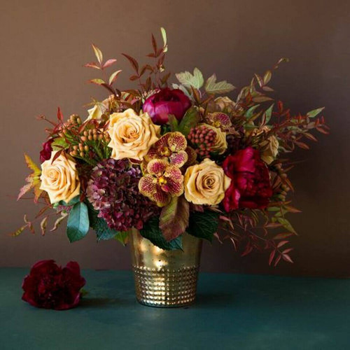 This beautiful floral arrangement offers a beautiful color maroon and honey glow. Rich burgundy peonies and cream colored roses contrast the rust hydrangea and orange vanda orchids placed throughout. Fall foliage ties everything together, with the blooms resting inside an 8” tall gold aluminum vase. Overall, this arrangement stands 17” tall.