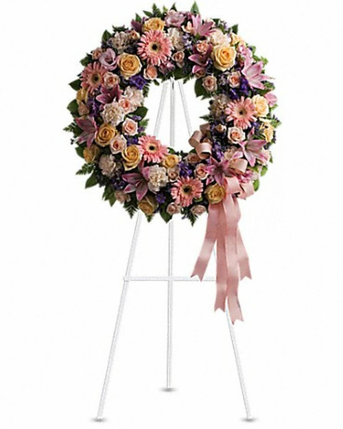 A mix of flowers such as peach roses, gerberas and carnations, pink asiatic lilies and lisianthus, purple limonium and lavender larkspur nest in greens on an easel-mounted wreath.