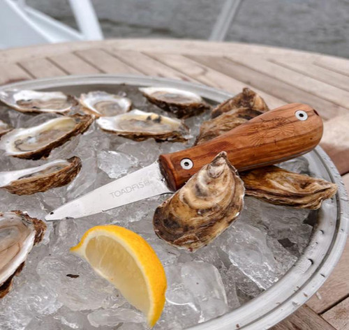 TOADFISH Toadfish Wood Handle Oyster Knife, a wooden-handled oyster shucking knife placed on a tray of fresh oysters with a lemon wedge, displayed on ice for seafood shucking.