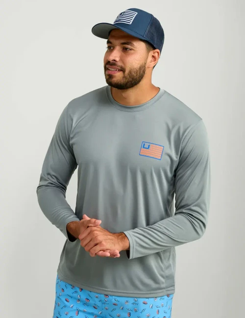 HUK Pursuit Huk And Bars LS Crew, man wearing a light gray long sleeve crew shirt with a U.S. flag graphic on the chest and a navy blue trucker hat with a striped patch