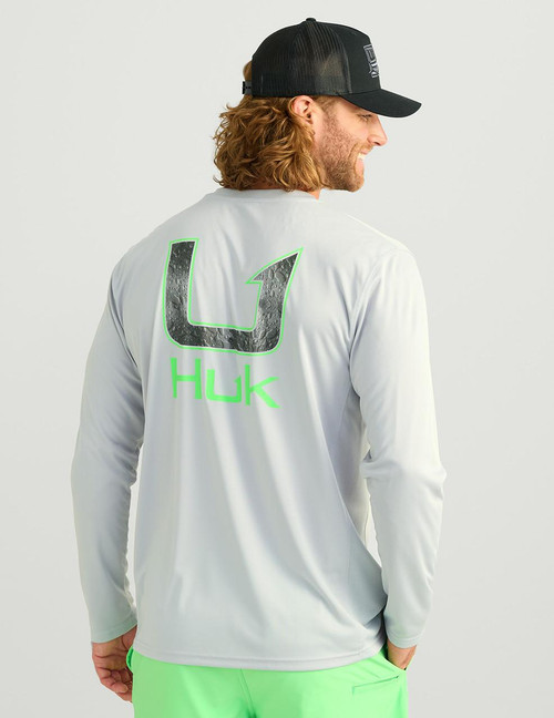 HUK Pursuit Huk Fill LS Crew, a man wearing a white long-sleeve athletic shirt with a large black and gray logo on the back and neon green detailing, against a plain gray background.
