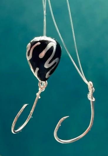 Hook'd On Reelin' Black Ice, close-up of a black and white fishing jig with two treble hooks and a black bead, blue background.