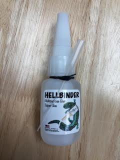 HELLBINDER Hellbinder Bait Glue, close-up of a white bottle with a pointed applicator tip and label featuring a green and black design on a wooden surface.
