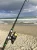 NINJA TACKLE Ninja Tackle 11' Dagger 2-7oz K Series Rod, fishing rod set up on a sandy beach with the ocean and cloudy sky in the background.
