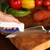 ACCUSHARP AccuSharp 001C Knife/Tool Sharpener, person using the sharpener on a kitchen knife, with fresh tomatoes and bell peppers on a wooden cutting board in the background.
