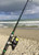 NINJA TACKLE Ninja Tackle 11' Dagger Spinning fishing rod, angled view on a sandy beach with ocean waves and cloudy sky in the background.