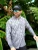 TORMENTER Tormenter SPF Hammerhead Camo Hoodie, a man wearing a gray camouflage hoodie with a fish skeleton logo, sunglasses, and a black cap, standing amidst lush green tropical plants.