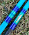 Nick's Custom Rods 2025, a fishing rod with a bright blue and green color scheme and a fish illustration on the handle, lying on grass.