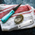TOADFISH Toadfish White Oyster Knife, stainless steel oyster shucking knives with ergonomic handles, one in white and one in turquoise, placed on a white and red striped cloth with an oyster shell.
