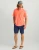HUK Kona Solid SS, man wearing a coral short-sleeve button-up shirt, navy shorts, a cap, and flip-flops.