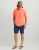 HUK Kona Solid SS, man wearing a coral short-sleeve button-up shirt, navy shorts, a trucker cap, and flip-flops.
